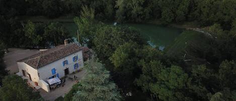 A white building with blue shutters, surrounded by dense greenery and a river.