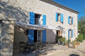 A stone building with blue shutters, a covered patio with chairs, and potted plants.