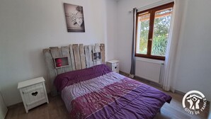 A bedroom with a wooden headboard, a bed with a purple and white bedspread, two bedside tables, and a window with a view of greenery. - La Bruninquillé - MONTREDON-LABEISSONNIE (MONTREDON-LABEISSONNIE)