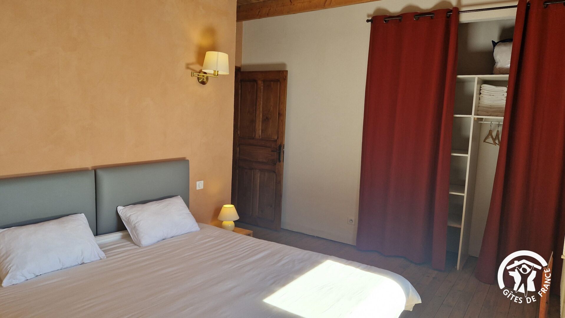 Une chambre à coucher avec un lit, deux oreillers, une table de chevet avec une lampe, une porte en bois et une armoire avec des rideaux rouges.
