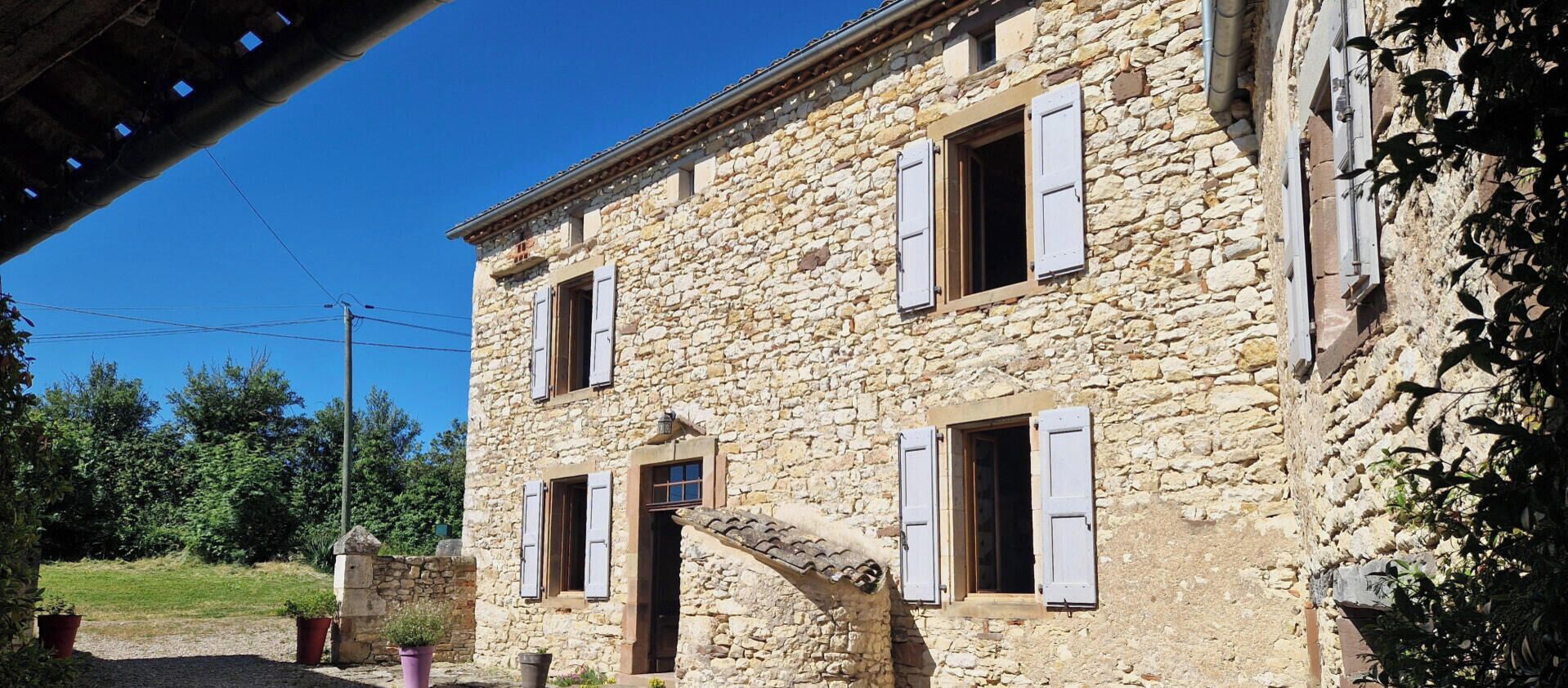A stone building with multiple windows and shutters, surrounded by gravel and potted plants.