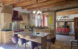 Cocina rústica con vigas de madera, una isla central con taburetes de bar, una pared de piedra y un comedor con mantel rojo.