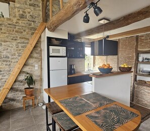 A rustic kitchen with exposed brick walls, wooden beams, a wooden dining table with patterned placemats, a built-in microwave, and a refrigerator.