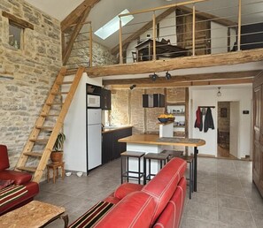 A rustic interior with a loft bedroom, stone walls, a kitchen with a wooden counter, and a red leather sofa.