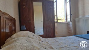 Een slaapkamer met een houten garderobe, een bed met een beddegoed met patroon, een bedlamp en een raam met uitzicht op bomen.