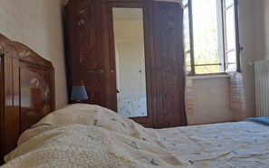 Une chambre avec une armoire en bois, un lit recouvert d’une couette à motifs, une lampe de chevet et une fenêtre donnant sur des arbres.