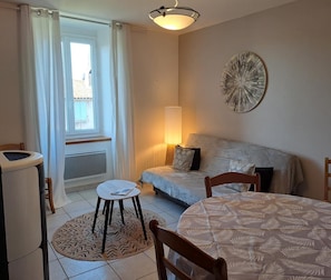 A cozy living room with a sofa, a round table, and a small stool. There is a window with curtains, a lamp, and a decorative wall piece. - Gîte communal Le Vintrou 2 (Le Vintrou)