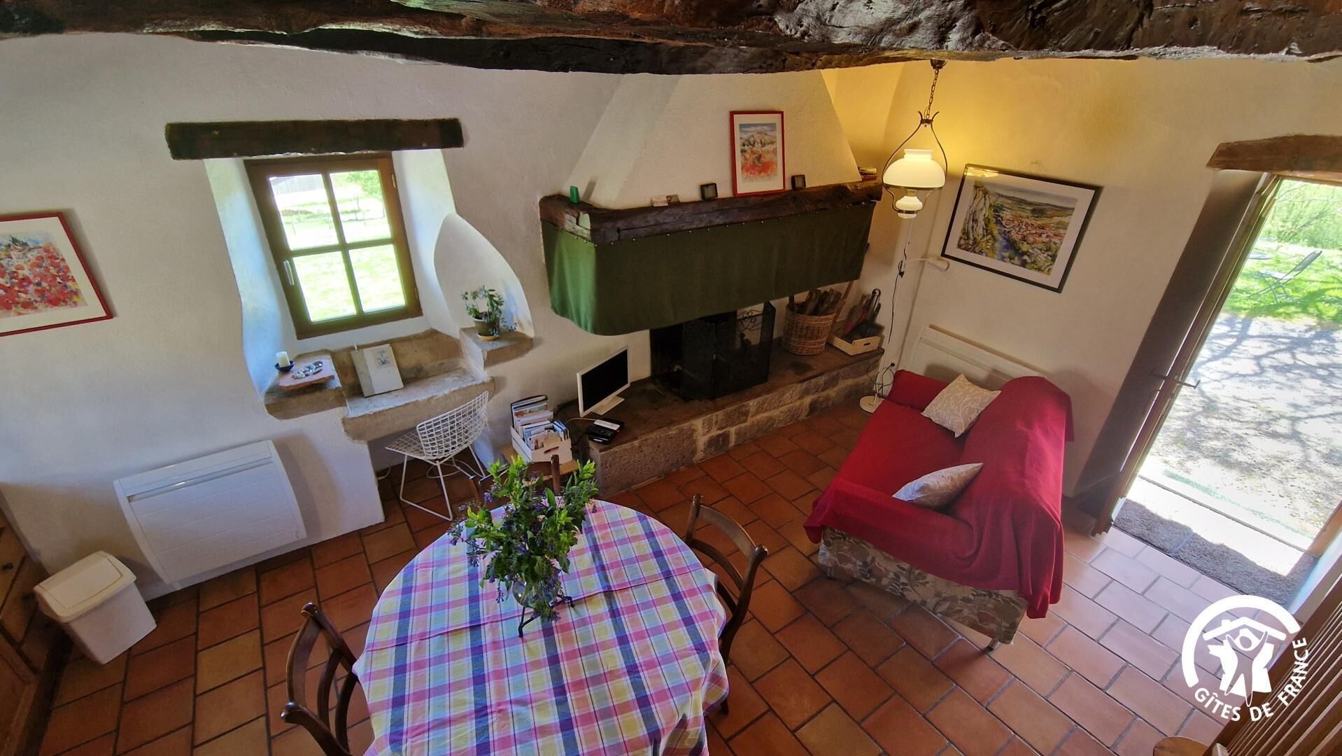 A cozy room with a fireplace, a red sofa, a dining table with a colorful tablecloth, and a window overlooking greenery.