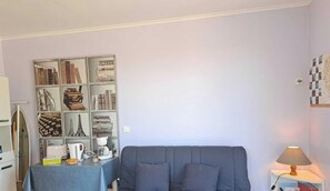 Ein Zimmer mit einem grauen Sofa, einem kleinen Tisch mit einer Lampe und einem Bücherregal mit verschiedenen Gegenständen.