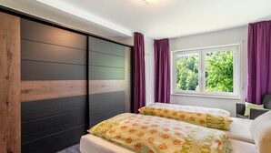 A bedroom with two beds, a window with curtains, a view of greenery, and a ceiling light fixture. - Apartment 'Burkhardt' with Mountain View, Private Terrace and Wi-Fi (Bad Herrenalb)