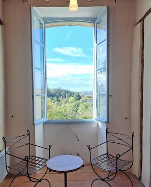 Ein Zimmer mit großem Fenster, zwei Metallstühlen, einem kleinen runden Tisch und einer Pendelleuchte.