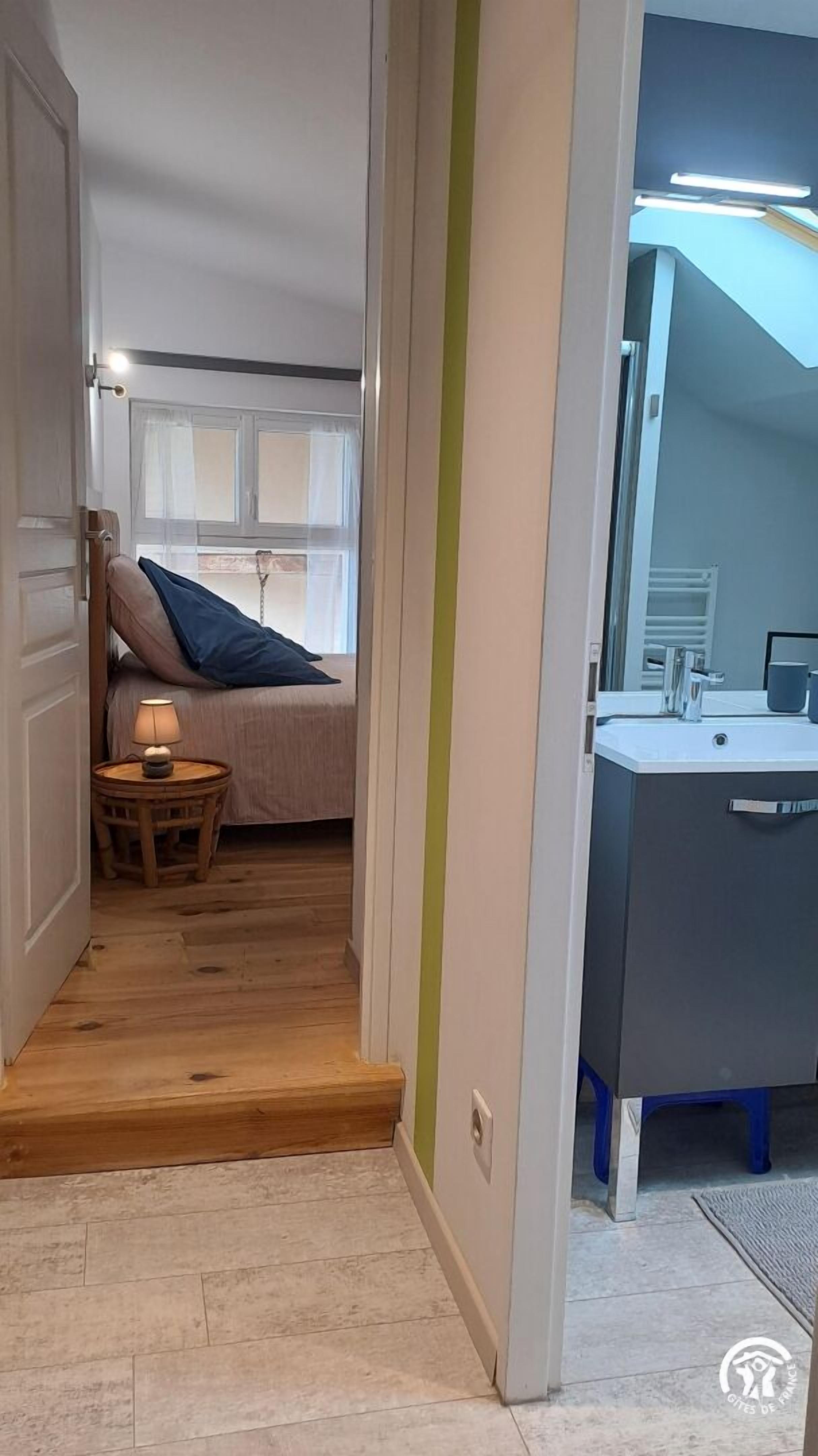 A narrow hallway with a wooden floor leading to a bedroom with a bed, a nightstand, and a window. A bathroom with a sink and mirror is visible to the right.