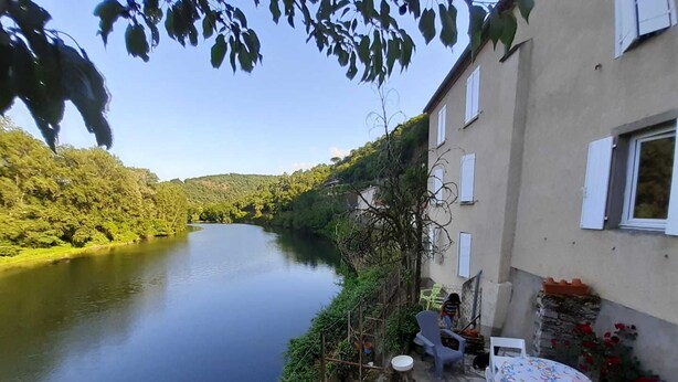 Un patio junto al río con suelo de piedra, mobiliario exterior y vistas a un río tranquilo.
