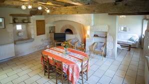 A rustic dining area with a tiled floor, a table with a floral tablecloth, wooden chairs, a stone fireplace, and a television. - La Falgarié - VIRAC - Gîte (VIRAC)