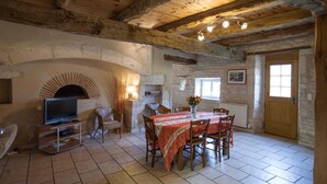 A rustic dining area with a tiled floor, a wooden table with chairs, a television on a stand, and a stone fireplace. - La Falgarié - VIRAC - Gîte (VIRAC)