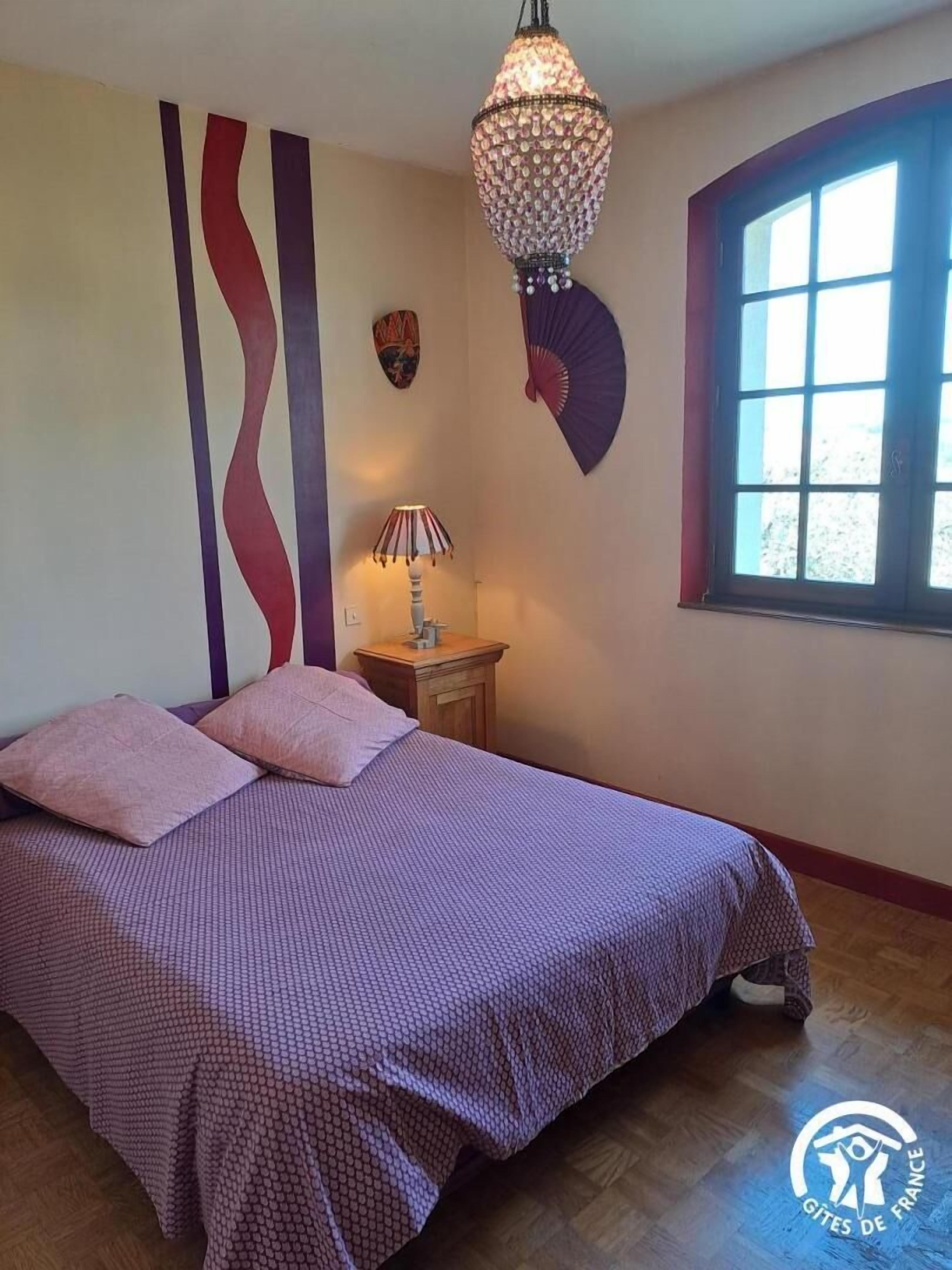 Une chambre à coucher comprenant un lit, une table de chevet, une lampe, un élément décoratif mural et une fenêtre.