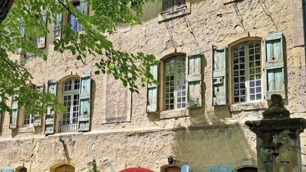 A building with multiple windows, a small outdoor seating area with a red umbrella, and a stone pillar. - The kitchens of Château d'Appelle (APPELLE)