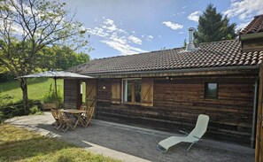 Une cabane en bois rustique, dotée d’un toit en tuiles, d’une terrasse avec une table et des chaises, et d’un fauteuil de relaxation.