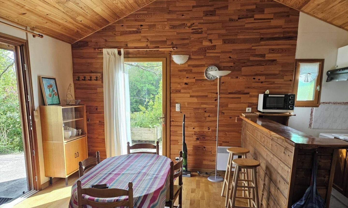 A cozy cabin with a wooden interior, a dining table with chairs, a kitchen area with a microwave, and a view of greenery outside.