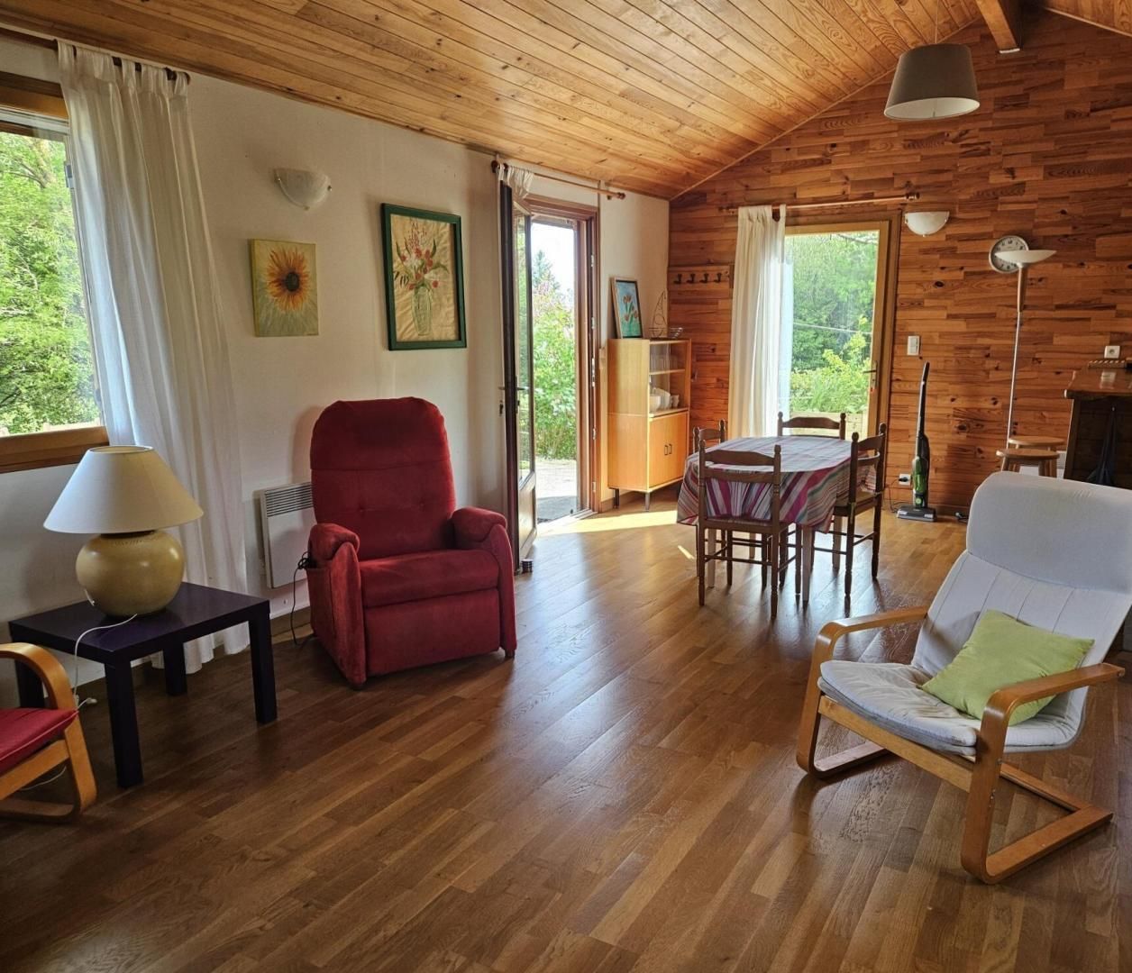 A cozy cabin with wooden walls and floors, a dining area with a table and chairs, a red armchair, a white armchair with a green pillow, and a lamp on a side table.