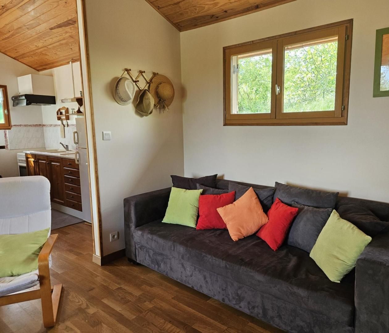 A cozy living area with a dark grey sofa, colorful cushions, and a wooden ceiling. A kitchen is visible in the background with a window overlooking greenery.