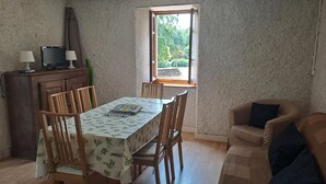 Kompakti oleskelutila, jossa on ruokapöytä, tuolit, sohva ja televisio, joka on sijoitettu puiseen kaappiin. Ikkunasta avautuu näkymä vehreään maisemaan.