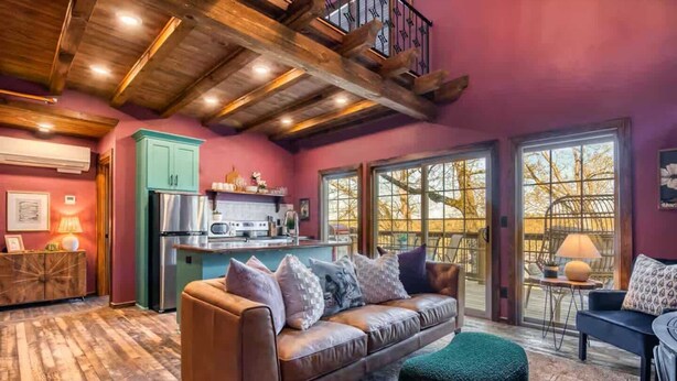 A cozy living room with a leather sofa, a green ottoman, and a dining area with a table and chairs. A kitchen with a refrigerator and a window is visible in the background. - Stay in the Tree House! One of a Kind Experience! (Branson)