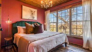 A bedroom with a large bed, a chandelier, a painting of a horse, and a window with a view of trees. - Stay in the Tree House! One of a Kind Experience! (Branson)