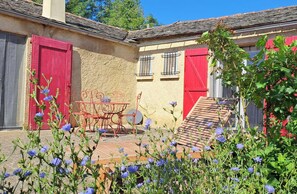 Un bĂątiment rustique avec une porte rouge, un volet rouge et un petit patio agrĂ©mentĂ© dâune table et de chaises.