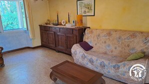 A living room with a patterned sofa, a wooden cabinet, a small table, and a window with curtains. - Au pré vert - SAINT PIERRE DE TRIVISY (SAINT PIERRE DE TRIVISY)