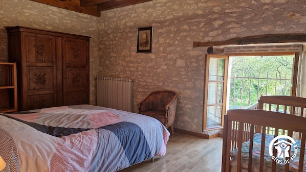 Une chambre à coucher rustique avec un mur en pierre, un lit en bois, une chaise et une fenêtre donnant sur un paysage verdoyant.