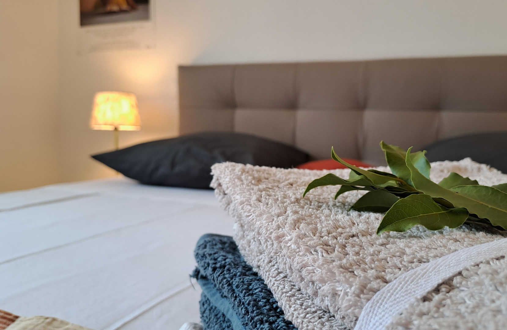 Ein ordentlich bezogenes Bett mit einer weiß-blauen Decke, einem grauen Kopfteil und einer dekorativen Anordnung von Lorbeerblättern.