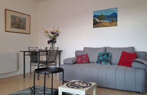 Ett vardagsrum med en grå soffa, ett litet runt bord, ett matbord med stolar och en vas med blommor. Det hänger två tavlor på väggen.