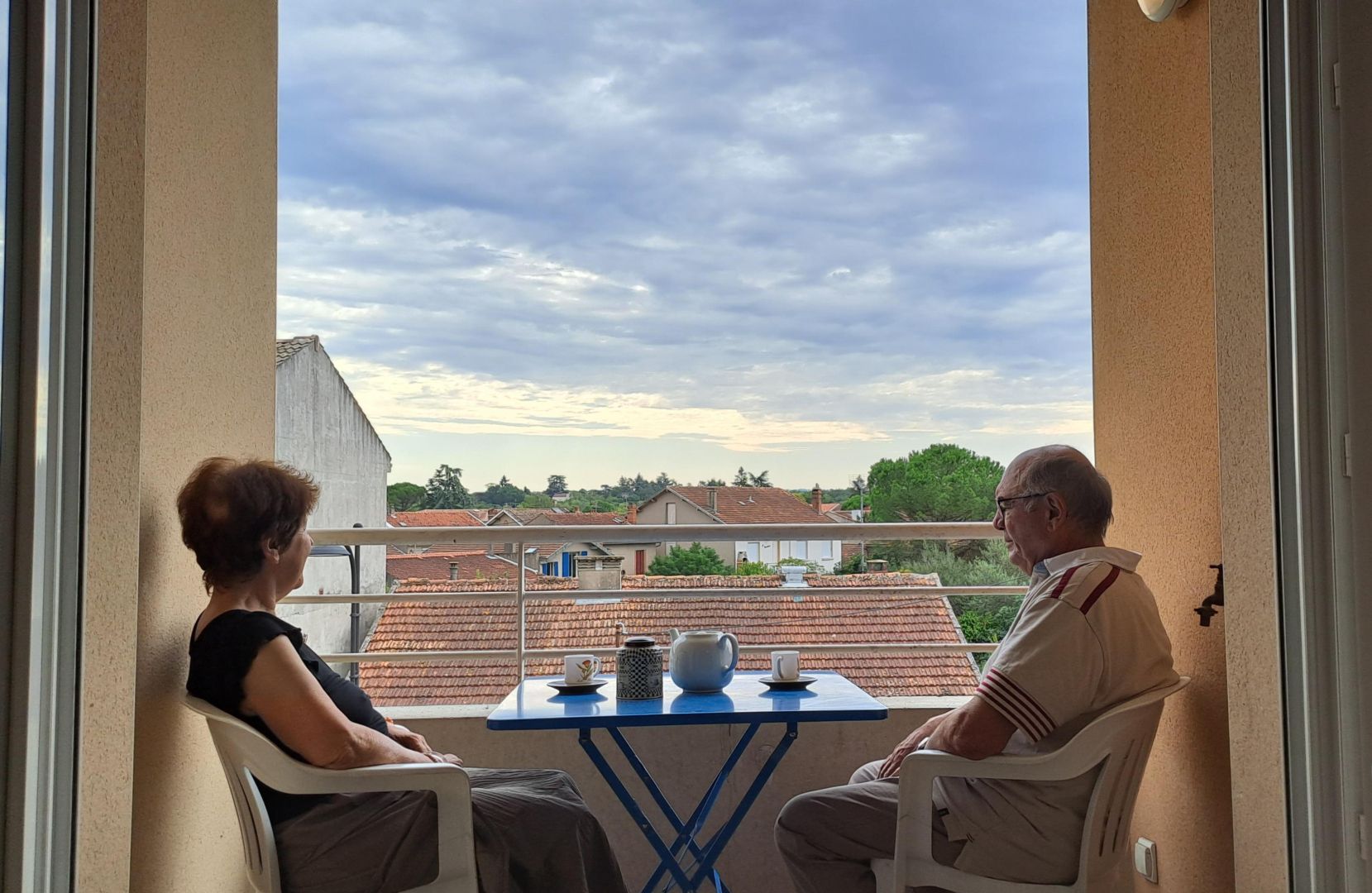 Un balcón con dos sillas, una mesa y vistas a un barrio residencial con casas y árboles.