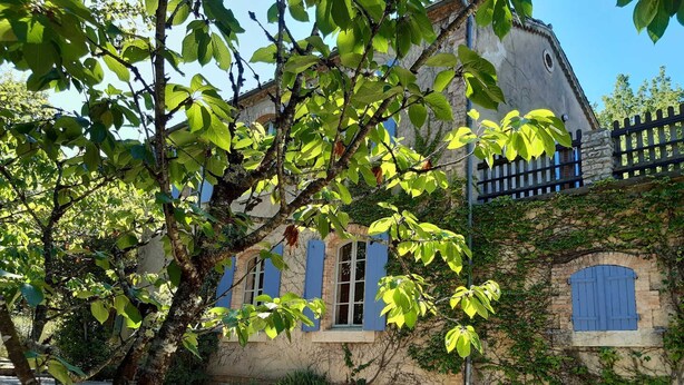 A stone building with blue shutters, surrounded by greenery and a stone wall.