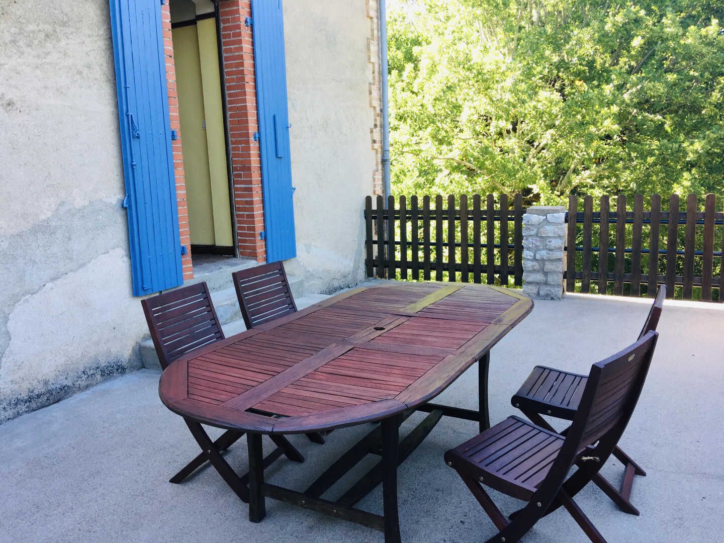 Un patio al aire libre con una mesa y sillas de madera, un muro de ladrillo con contraventanas azules y una valla de madera.