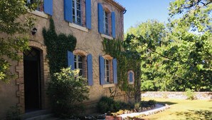A rustic stone building with blue shutters, a wooden table and chairs, and a garden with trees and a stone wall.