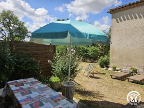 Een terras met een tafel, afgedekt met een tafelkleed met bloemenprint, een blauwe parasol en een houten omheining.