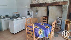 Une cuisine compacte avec des meubles blancs, un four à micro-ondes et une cuisinière. On y trouve un coin repas avec une table recouverte d’une nappe à motifs de tournesols et des chaises en bois.
