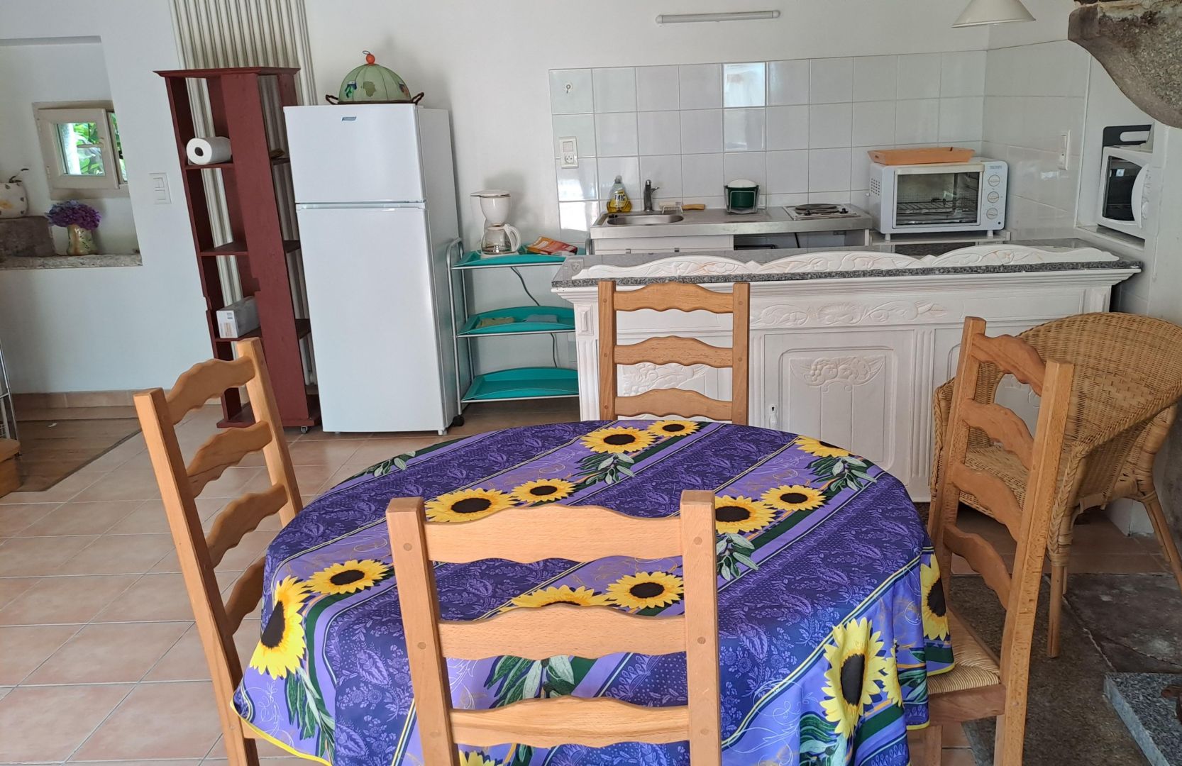 A kitchen with a dining table covered by a sunflower tablecloth, wooden chairs, a refrigerator, a microwave, and a sink.