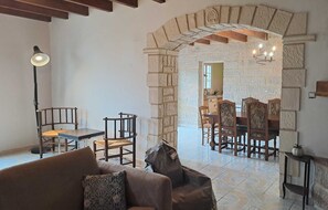 A cozy living area with a stone archway leading to a dining room. There is a sofa, a table, and chairs in the living area, and a chandelier above the dining table.