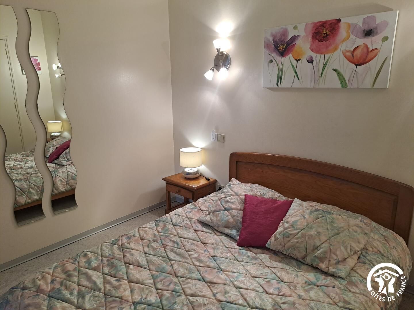 A bedroom with a bed, a nightstand, a lamp, a mirror, and a floral painting on the wall.
