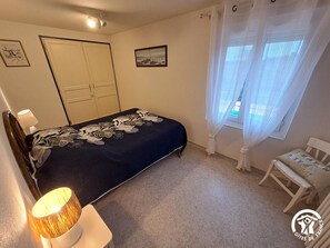 Um quarto com cama, cadeira, luminária, guarda-roupa e janela com cortinas.