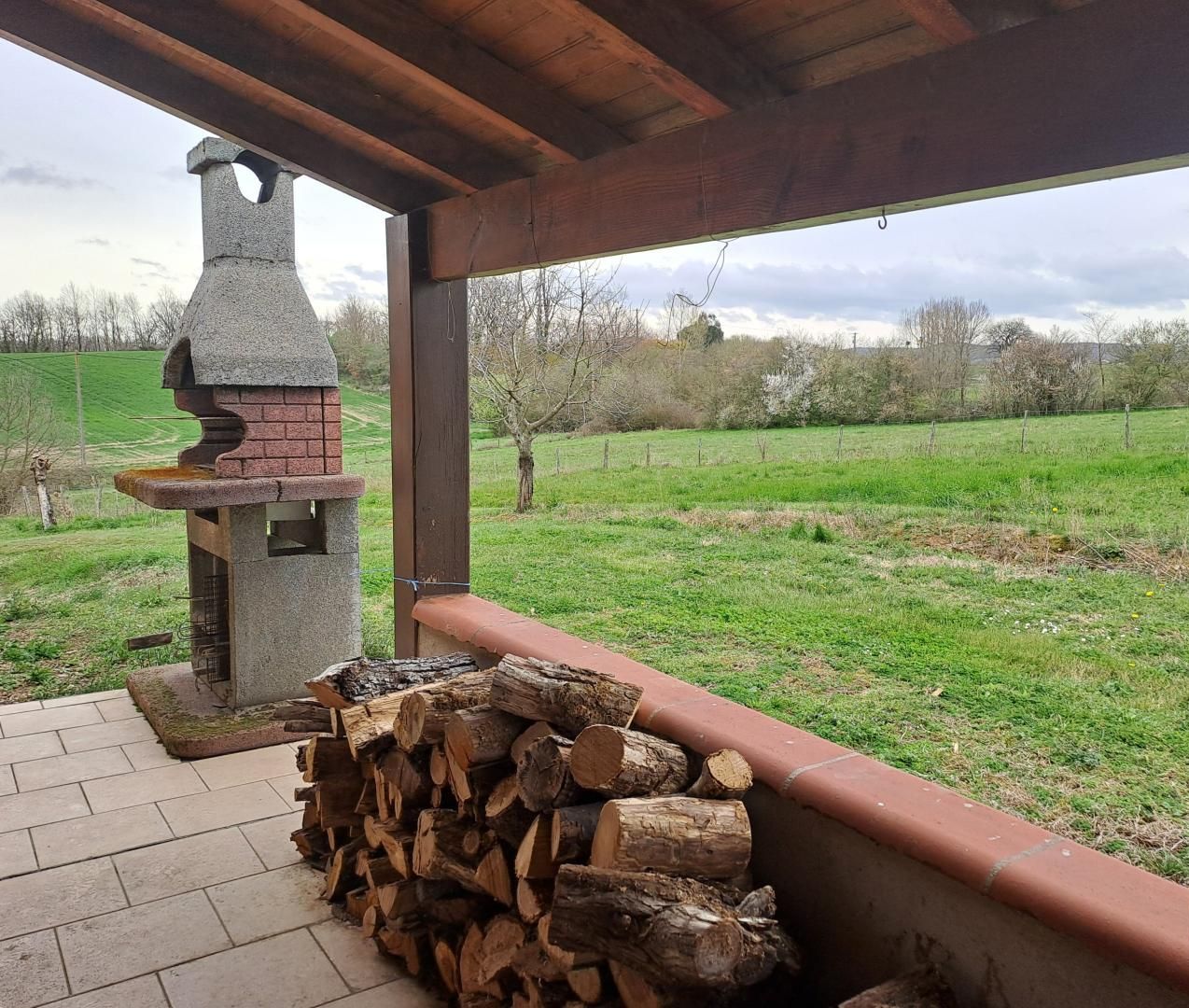 Ein rustikaler Außenbereich mit einem Steinofen, einem Holzstapel und Blick auf ein grünes Feld.