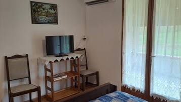 Une chambre à coucher avec un lit, un téléviseur posé sur une petite table, deux chaises, une fenêtre avec des rideaux et un tableau accroché au mur.