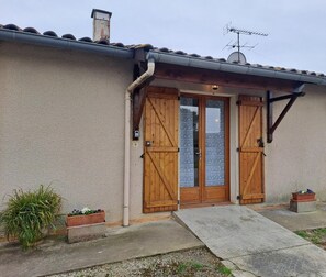 Ein Haus mit einer beige gestrichenen Fassade, einem Ziegeldach und einem Schornstein. Es gibt zwei Holztüren mit Glasfenstern, eine kleine Veranda mit einer Betonstufe und eine Antenne auf dem Dach.