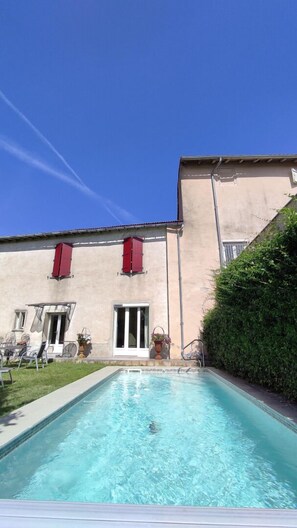 A swimming pool with a clear blue water, a building with red shutters, and a hedge on the right. - Le relais de la poste (CASTELNAU DE MONTMIRAL)