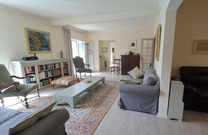 Un salon avec un canapé, un fauteuil et une table basse. On aperçoit une bibliothèque et un meuble en arrière-plan.