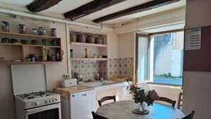 A kitchen with a gas stove, oven, and washing machine. There is a dining table with chairs and a vase of flowers. Shelves with jars and kitchen items are visible. - Les Bons Enfants - VENES (VENES)