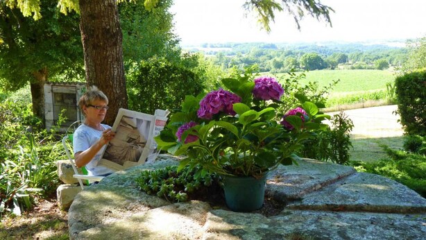A person reading a newspaper in a garden with a stone bench, surrounded by greenery and a view of a landscape. - Saint Martial - Terre de Bancalié (Terre de Bancalié)